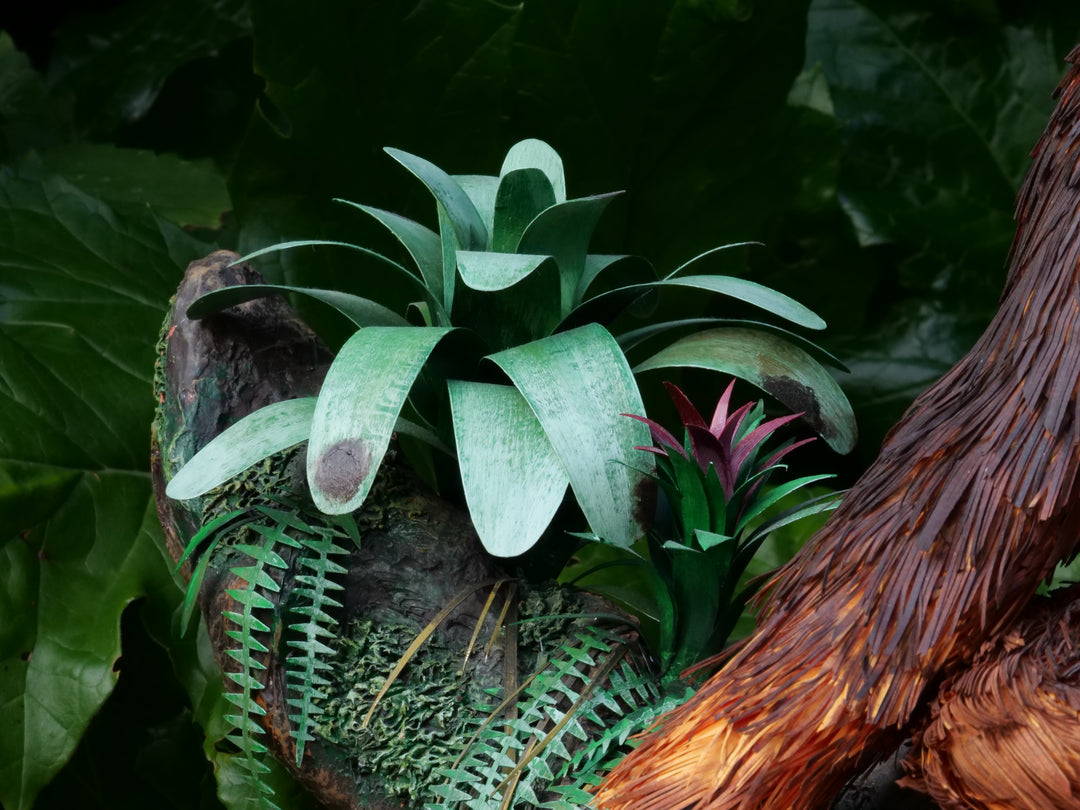 Highly detailed sculpture of a Bornean orangutan lounging on a branch, featuring hand-cut burnt orange paper fur, textured grey-blue skin, and expressive amber eyes, surrounded by lush green tropical foliage.