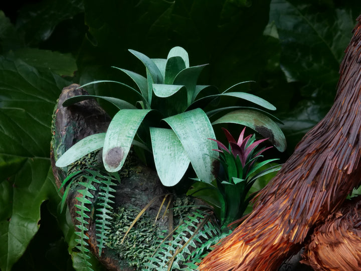 Highly detailed sculpture of a Bornean orangutan lounging on a branch, featuring hand-cut burnt orange paper fur, textured grey-blue skin, and expressive amber eyes, surrounded by lush green tropical foliage.