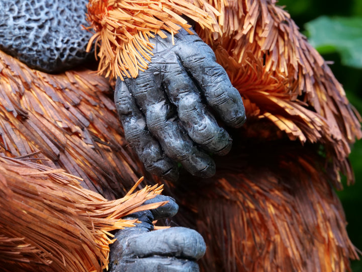 Highly detailed sculpture of a Bornean orangutan lounging on a branch, featuring hand-cut burnt orange paper fur, textured grey-blue skin, and expressive amber eyes, surrounded by lush green tropical foliage.
