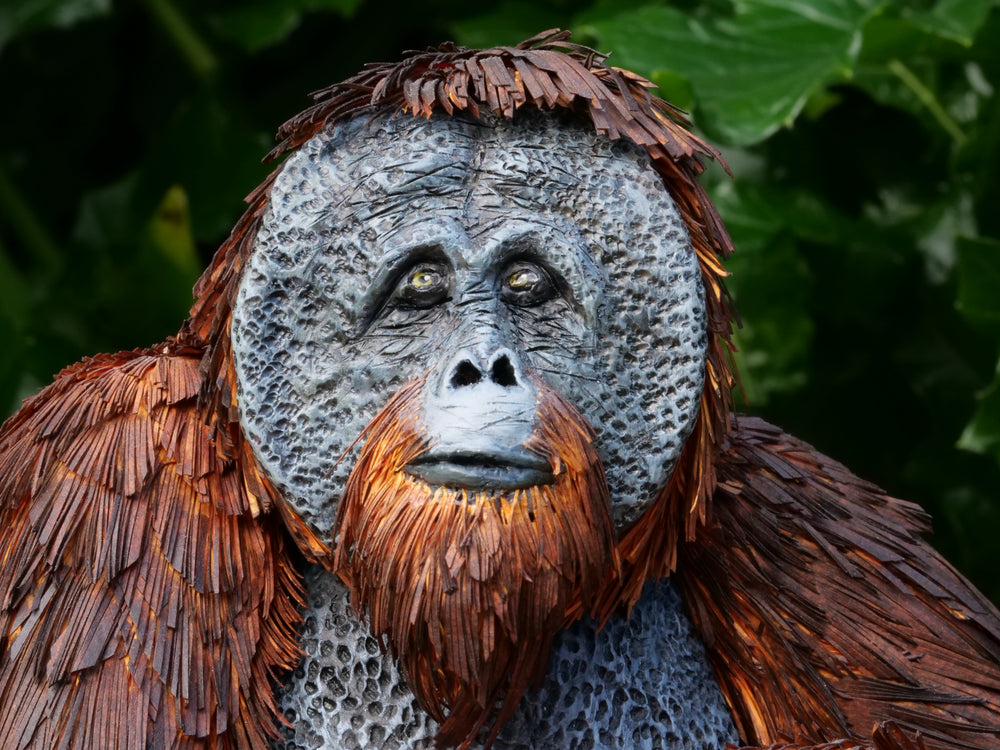 Highly detailed sculpture of a Bornean orangutan lounging on a branch, featuring hand-cut burnt orange paper fur, textured grey-blue skin, and expressive amber eyes, surrounded by lush green tropical foliage.