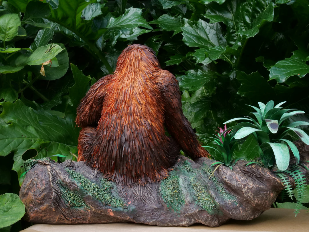 Highly detailed sculpture of a Bornean orangutan lounging on a branch, featuring hand-cut burnt orange paper fur, textured grey-blue skin, and expressive amber eyes, surrounded by lush green tropical foliage.