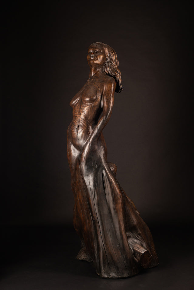 Tall figurative sculpture of a woman carved from richly grained wood, standing with her head lifted toward the sky. Her form emerges organically from the trunk, evoking growth and connection to the earth. The flowing grain patterns enhance her posture, while a small child figure is carved into the back of the sculpture, reaching upward and held by the woman's hand. A deeply symbolic piece that blends maternal strength, natural cycles, and generational continuity. The bronze preserves the texture of wood.