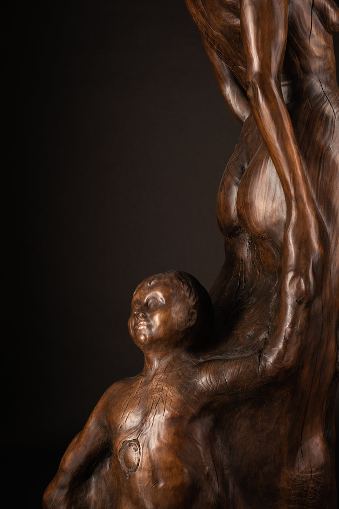 Tall figurative sculpture of a woman carved from richly grained wood, standing with her head lifted toward the sky. Her form emerges organically from the trunk, evoking growth and connection to the earth. The flowing grain patterns enhance her posture, while a small child figure is carved into the back of the sculpture, reaching upward and held by the woman's hand. A deeply symbolic piece that blends maternal strength, natural cycles, and generational continuity. The bronze preserves the texture of wood.