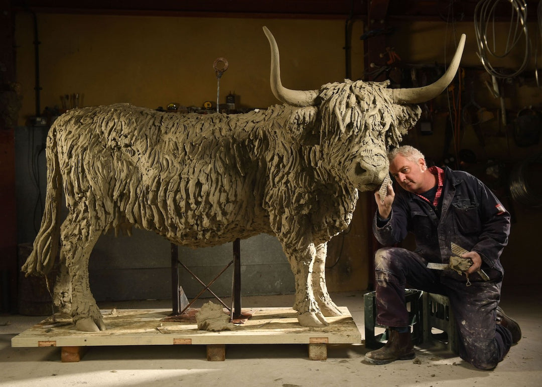 ‘Highland Cow: Life Size’ by Hamish Mackie, a life-size limited-edition bronze sculpture depicting a Scottish Highland cow standing in a grassy field. The sculpture features long, sweeping horns, a broad muzzle and a heavily textured, shaggy coat falling over the animal’s eyes. Finished in warm earthy brown bronze, the realistic wildlife sculpture captures the distinctive character and rugged appearance of the Highland breed.
