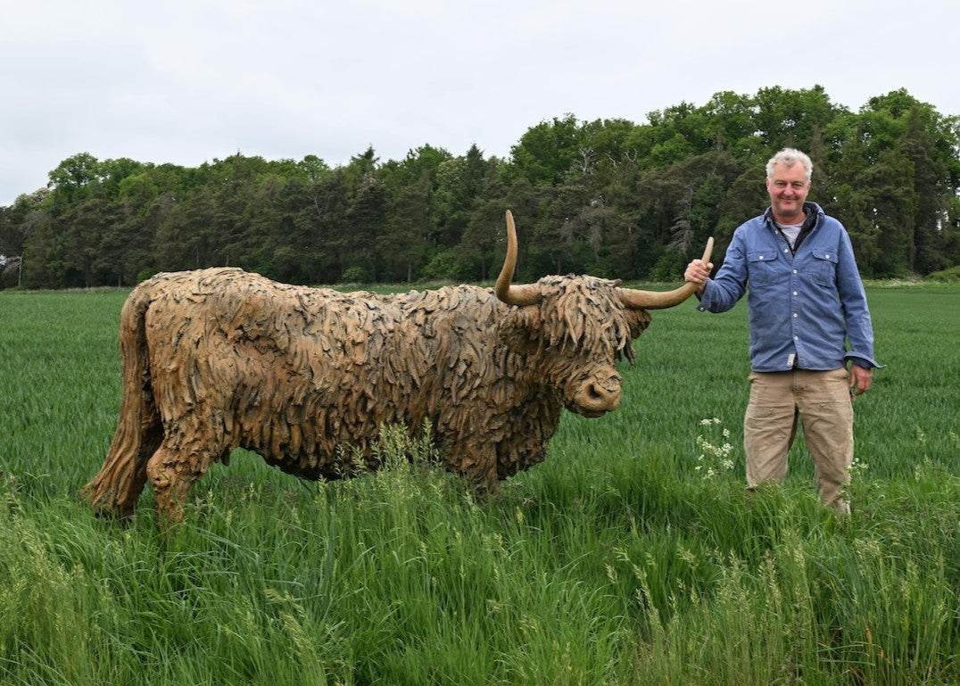 ‘Highland Cow: Life Size’ by Hamish Mackie, a life-size limited-edition bronze sculpture depicting a Scottish Highland cow standing in a grassy field. The sculpture features long, sweeping horns, a broad muzzle and a heavily textured, shaggy coat falling over the animal’s eyes. Finished in warm earthy brown bronze, the realistic wildlife sculpture captures the distinctive character and rugged appearance of the Highland breed.