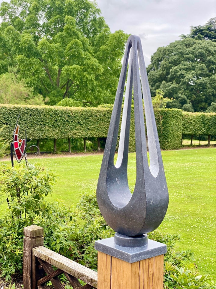Abstract sculpture Maja – 2024 by Hamish Holman, carved from dark Kilkenny limestone into a tall, teardrop-like form with elegant open voids and flowing symmetry, mounted on a stone base and oak plinth, designed for indoor or outdoor display.