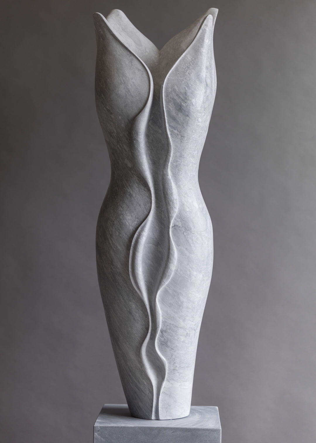 Tall contemporary sculpture hand-carved from pale Italian marble, featuring an elegant elongated form with flowing, organic curves and soft wave-like lines, poised on a smooth stone plinth with a serene, modern presence.