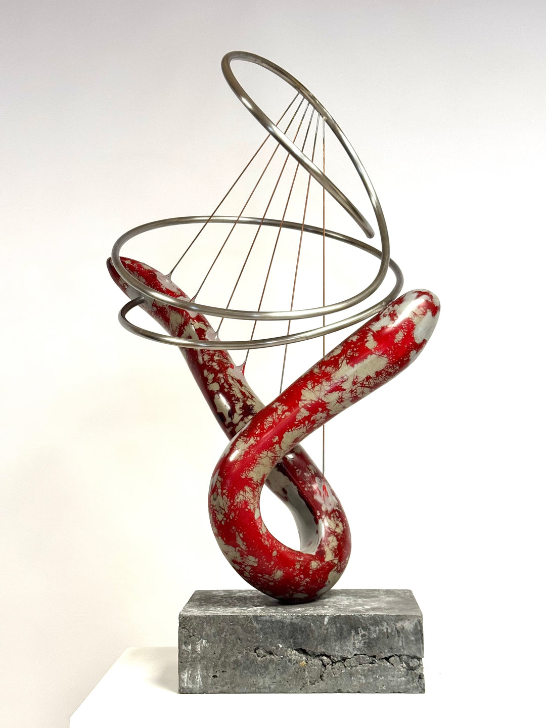 Abstract mixed media sculpture with looping red and silver marbled GRP form, intersected by brushed stainless steel rings and fine rust-treated steel strands, mounted on a grey stone base. Contemporary one-off artwork by Mark Purllant.