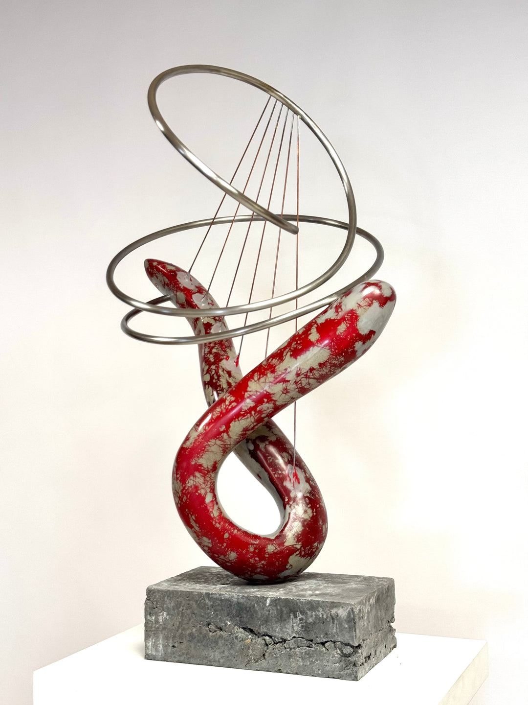 Abstract mixed media sculpture with looping red and silver marbled GRP form, intersected by brushed stainless steel rings and fine rust-treated steel strands, mounted on a grey stone base. Contemporary one-off artwork by Mark Purllant.