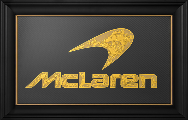 ‘McLaren Carbon’ by Vince Edmunds, a handcrafted Formula One automotive artwork featuring the iconic McLaren logo recreated in 24-carat gold leaf and glue-chipped glass. The distinctive McLaren speedmark and lettering feature intricate, individually formed gold textures against a dark charcoal-grey carbon fibre background with a visible diagonal weave. Presented in a wide satin black frame with a fine metallic gold sight line, combining British motorsport heritage with traditional glass gilding and modern c