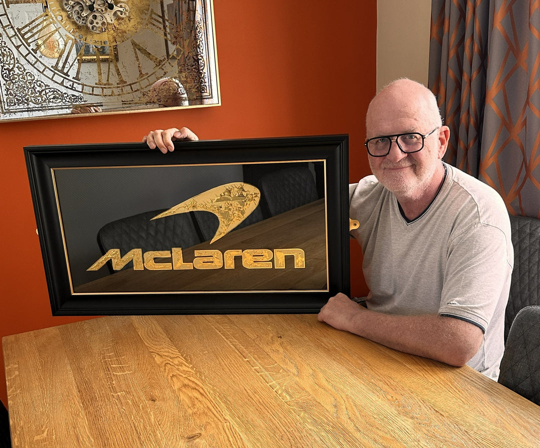 ‘McLaren Carbon’ by Vince Edmunds, a handcrafted Formula One automotive artwork featuring the iconic McLaren logo recreated in 24-carat gold leaf and glue-chipped glass. The distinctive McLaren speedmark and lettering feature intricate, individually formed gold textures against a dark charcoal-grey carbon fibre background with a visible diagonal weave. Presented in a wide satin black frame with a fine metallic gold sight line, combining British motorsport heritage with traditional glass gilding and modern c