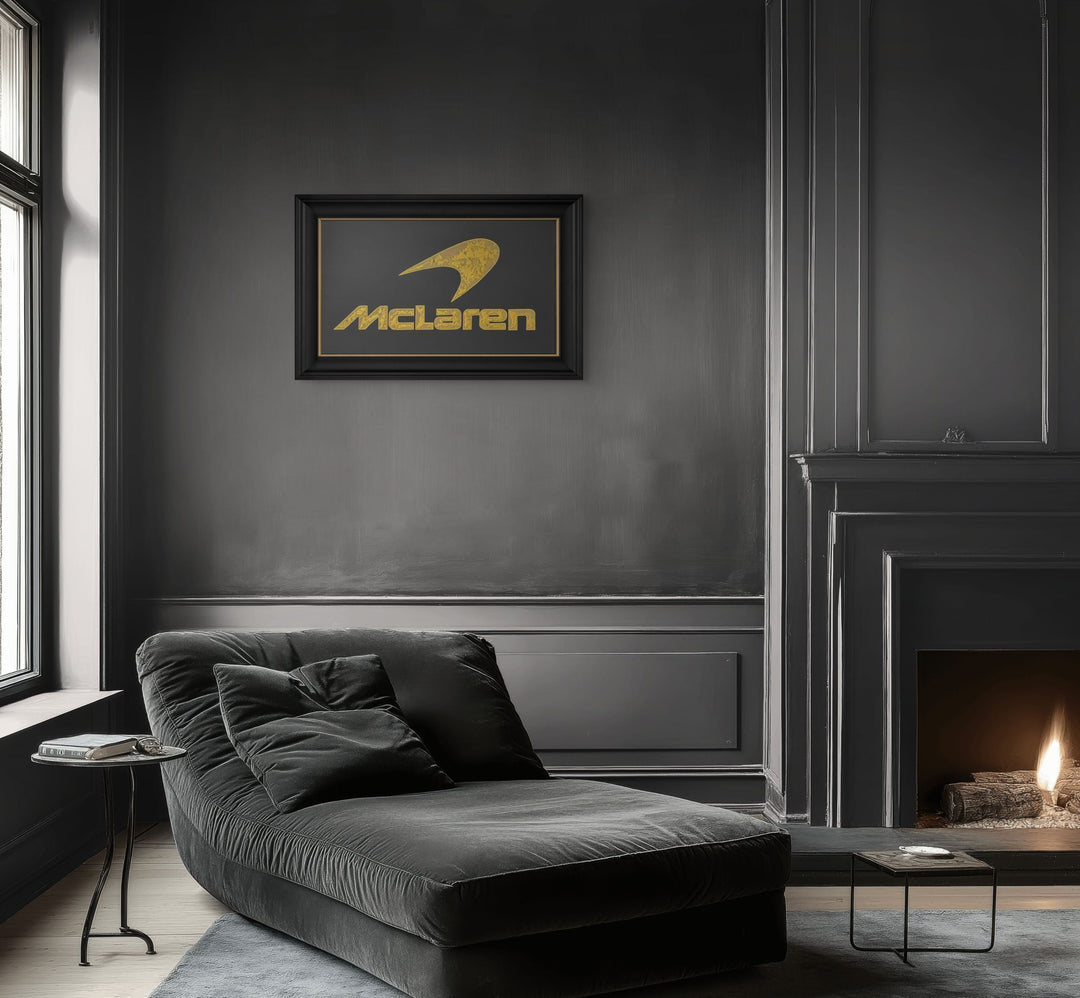 ‘McLaren Carbon’ by Vince Edmunds, a handcrafted Formula One automotive artwork featuring the iconic McLaren logo recreated in 24-carat gold leaf and glue-chipped glass. The distinctive McLaren speedmark and lettering feature intricate, individually formed gold textures against a dark charcoal-grey carbon fibre background with a visible diagonal weave. Presented in a wide satin black frame with a fine metallic gold sight line, combining British motorsport heritage with traditional glass gilding and modern c