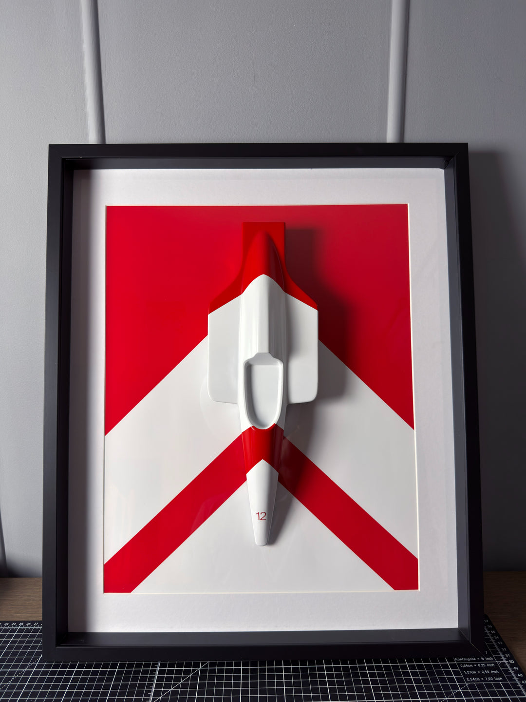‘McLaren MP4/4 Inspired Wall Art’ by Srini Gandra, a limited-edition contemporary automotive wall sculpture from the ICONS on The Wall series, celebrating Ayrton Senna and the legendary McLaren MP4/4 Formula 1 racing car. Handcrafted from resin and finished in high-gloss automotive paint, the three-dimensional sculpture features a streamlined white racing car viewed from above, with red detailing and the number 12 on its nose. Mounted against a bold red and white geometric background within a black wooden d