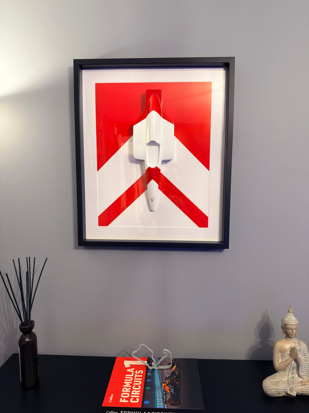 ‘McLaren MP4/4 Inspired Wall Art’ by Srini Gandra, a limited-edition contemporary automotive wall sculpture from the ICONS on The Wall series, celebrating Ayrton Senna and the legendary McLaren MP4/4 Formula 1 racing car. Handcrafted from resin and finished in high-gloss automotive paint, the three-dimensional sculpture features a streamlined white racing car viewed from above, with red detailing and the number 12 on its nose. Mounted against a bold red and white geometric background within a black wooden d
