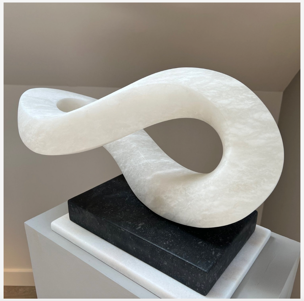 Abstract alabaster sculpture Meander – 2024 by Hamish Holman, hand-carved from Spanish alabaster into a fluid looping infinity-like form with soft translucent white tones, displayed on a contrasting dark stone base.