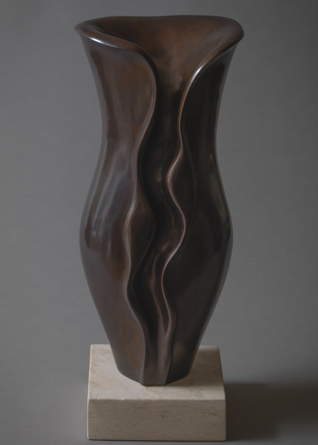 Sculptural bronze vessel with fluid, undulating contours that twist and fold organically upward, finished in a rich deep brown patina and set on a pale stone base.