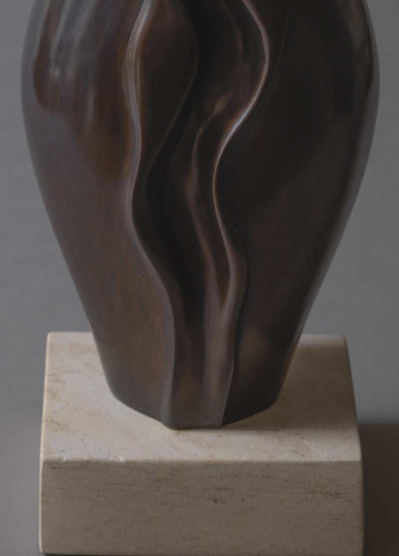 Sculptural bronze vessel with fluid, undulating contours that twist and fold organically upward, finished in a rich deep brown patina and set on a pale stone base.