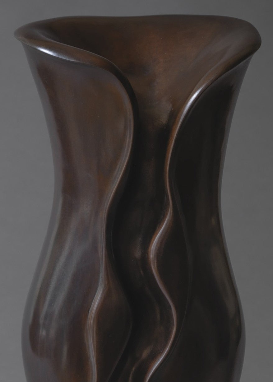 Sculptural bronze vessel with fluid, undulating contours that twist and fold organically upward, finished in a rich deep brown patina and set on a pale stone base.