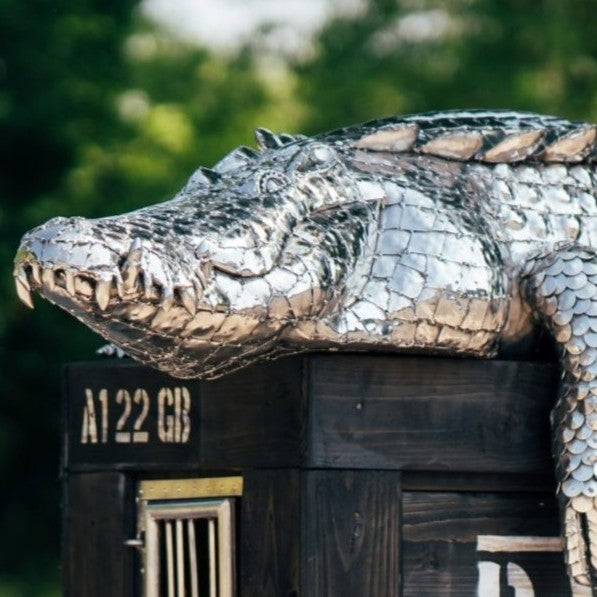‘Crocodile on a Box’ by Michael Turner, a unique, one-of-one stainless steel wildlife sculpture depicting a life-size crocodile resting across a large wooden crate. Handcrafted from intricately shaped and welded stainless steel, the crocodile features individually detailed silver scales, powerful jaws, raised ridges along its back, muscular legs and a long tail extending over the edge. The rustic brown timber box features a metal hatch, locking mechanism and bold black DANGER lettering, creating a striking 