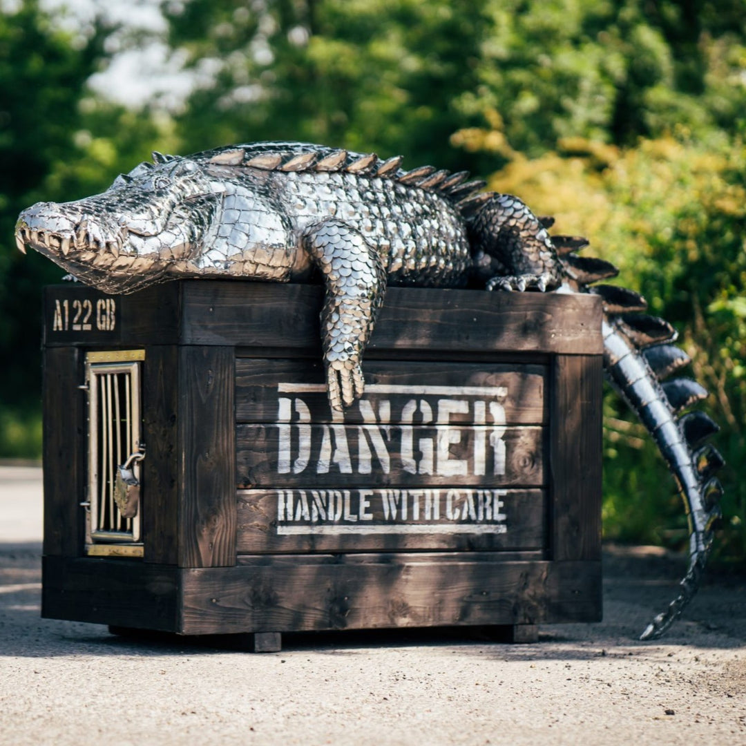 ‘Crocodile on a Box’ by Michael Turner, a unique, one-of-one stainless steel wildlife sculpture depicting a life-size crocodile resting across a large wooden crate. Handcrafted from intricately shaped and welded stainless steel, the crocodile features individually detailed silver scales, powerful jaws, raised ridges along its back, muscular legs and a long tail extending over the edge. The rustic brown timber box features a metal hatch, locking mechanism and bold black DANGER lettering, creating a striking 