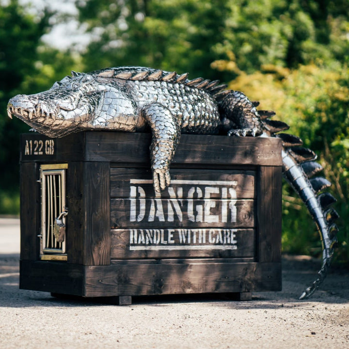 ‘Crocodile on a Box’ by Michael Turner, a unique, one-of-one stainless steel wildlife sculpture depicting a life-size crocodile resting across a large wooden crate. Handcrafted from intricately shaped and welded stainless steel, the crocodile features individually detailed silver scales, powerful jaws, raised ridges along its back, muscular legs and a long tail extending over the edge. The rustic brown timber box features a metal hatch, locking mechanism and bold black DANGER lettering, creating a striking 