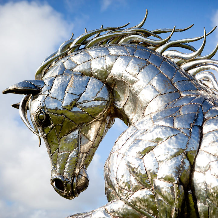 ‘Leaping Horse’ by Michael Turner, a unique, one-of-one stainless steel equestrian sculpture depicting a magnificent life-size horse leaping forward with its front legs raised and hind legs extended. Handcrafted from marine-grade stainless steel, the sculpture features intricately welded metallic plates, a powerful muscular body, flowing mane and sweeping tail. Its polished silver finish reflects the surrounding landscape, while the dynamic pose captures the strength, elegance and movement of a horse in ful