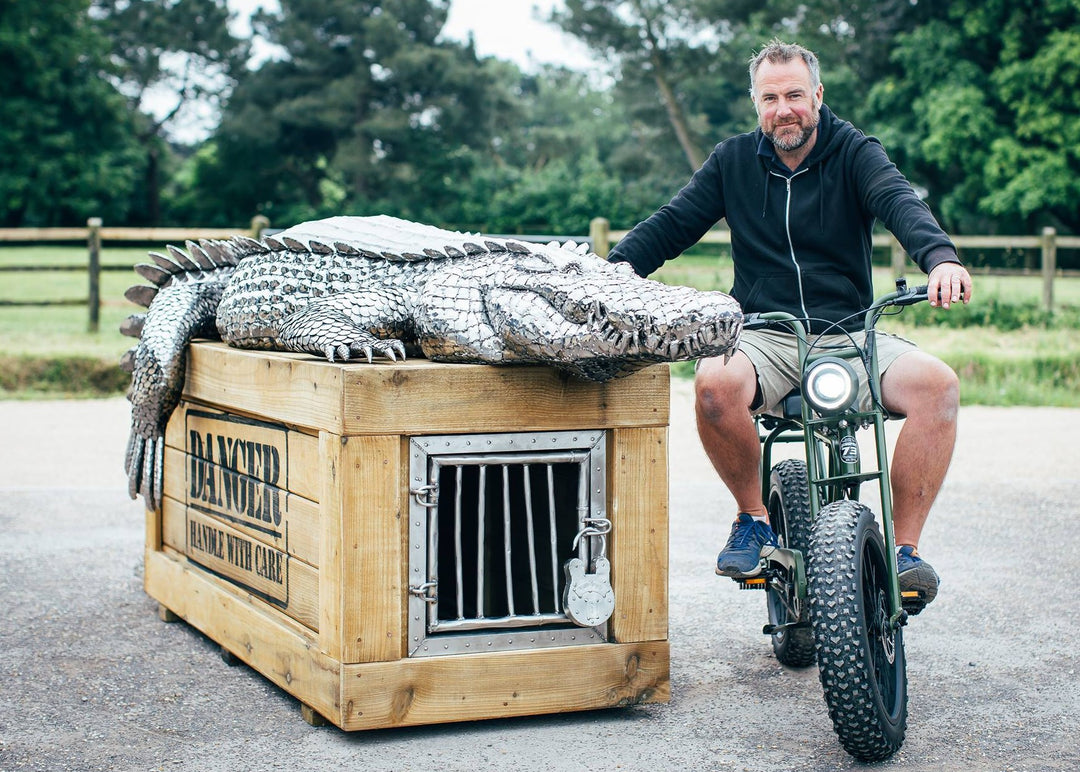 ‘Crocodile on a Box’ by Michael Turner, a unique, one-of-one stainless steel wildlife sculpture depicting a life-size crocodile resting across a large wooden crate. Handcrafted from intricately shaped and welded stainless steel, the crocodile features individually detailed silver scales, powerful jaws, raised ridges along its back, muscular legs and a long tail extending over the edge. The rustic brown timber box features a metal hatch, locking mechanism and bold black DANGER lettering, creating a striking 