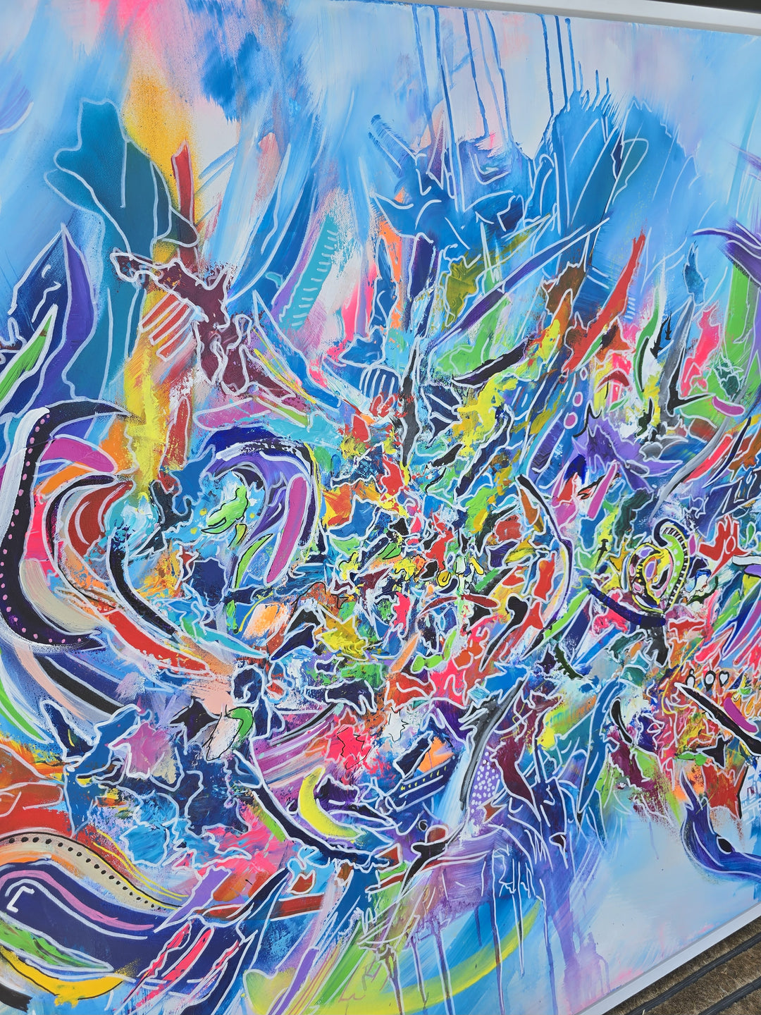 Vibrant large-scale abstract graffiti painting bursting with layered colour, bold sweeping lines and dynamic shapes against a bright blue background, framed in a white tray frame.