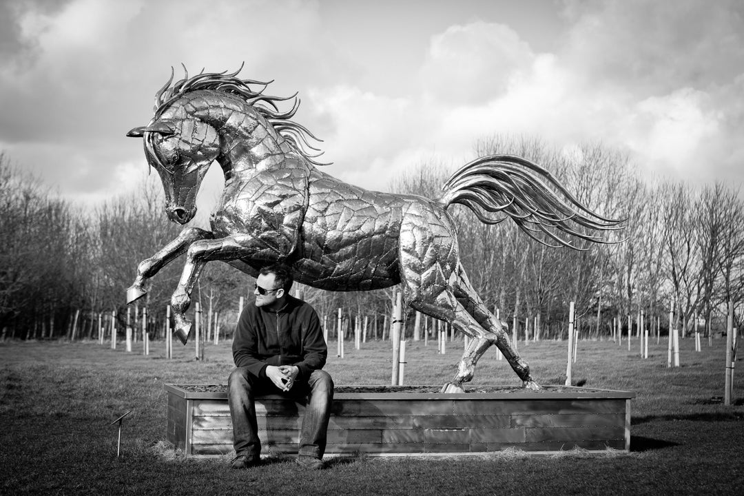 ‘Leaping Horse’ by Michael Turner, a unique, one-of-one stainless steel equestrian sculpture depicting a magnificent life-size horse leaping forward with its front legs raised and hind legs extended. Handcrafted from marine-grade stainless steel, the sculpture features intricately welded metallic plates, a powerful muscular body, flowing mane and sweeping tail. Its polished silver finish reflects the surrounding landscape, while the dynamic pose captures the strength, elegance and movement of a horse in ful