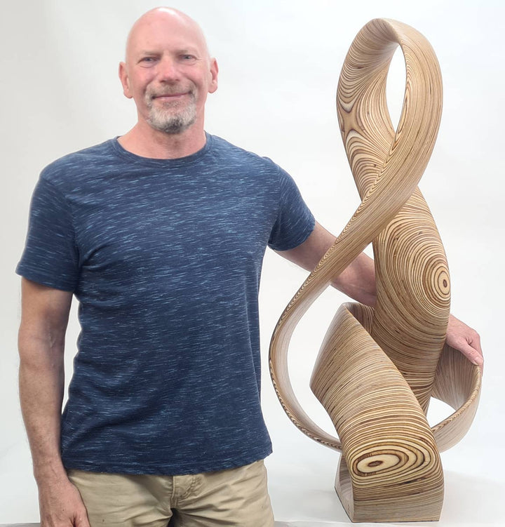 Hand-carved birch plywood sculpture by Bill Prickett combining a treble clef and Möbius strip into a continuous, flowing form, with layered wood grain revealing an infinity loop inspired by music and mathematics.