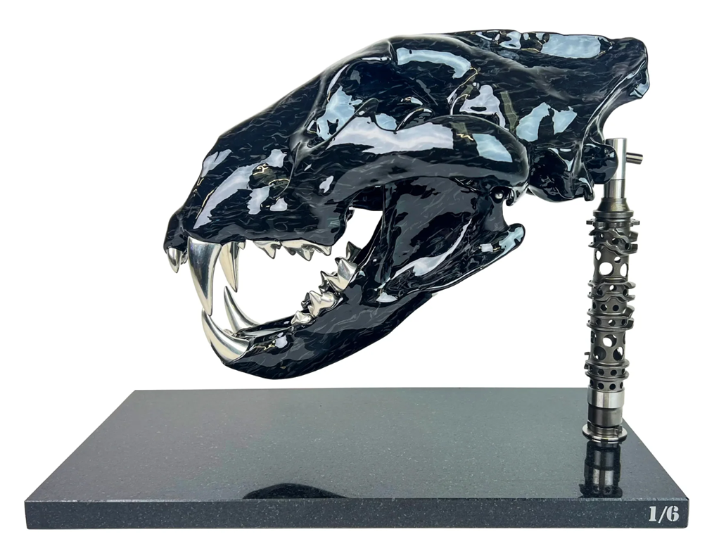 A striking, polished carbon fibre lion skull sculpture by Alastair Gibson, crafted with Mokume-style carbon layup for a swirling, chaotic texture. The jaw is fitted with gleaming bismuth alloy teeth. Mounted on a genuine F1 gearbox selector barrel atop a granite base, the work merges natural form with elite motorsport engineering.