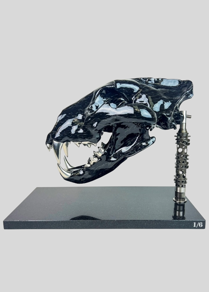A striking, polished carbon fibre lion skull sculpture by Alastair Gibson, crafted with Mokume-style carbon layup for a swirling, chaotic texture. The jaw is fitted with gleaming bismuth alloy teeth. Mounted on a genuine F1 gearbox selector barrel atop a granite base, the work merges natural form with elite motorsport engineering.