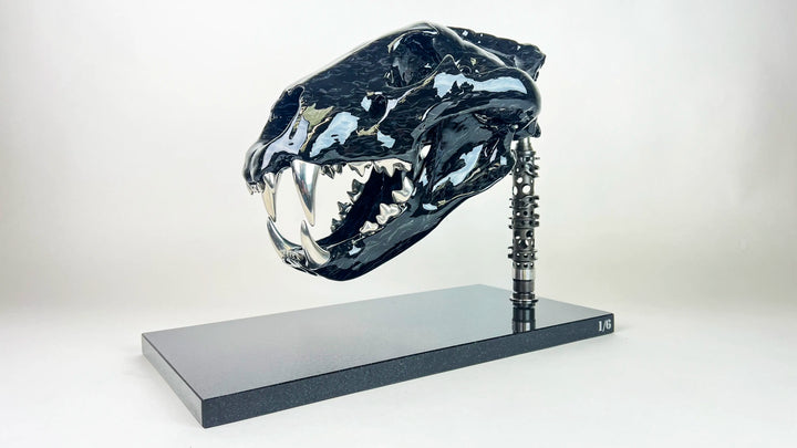 A striking, polished carbon fibre lion skull sculpture by Alastair Gibson, crafted with Mokume-style carbon layup for a swirling, chaotic texture. The jaw is fitted with gleaming bismuth alloy teeth. Mounted on a genuine F1 gearbox selector barrel atop a granite base, the work merges natural form with elite motorsport engineering.