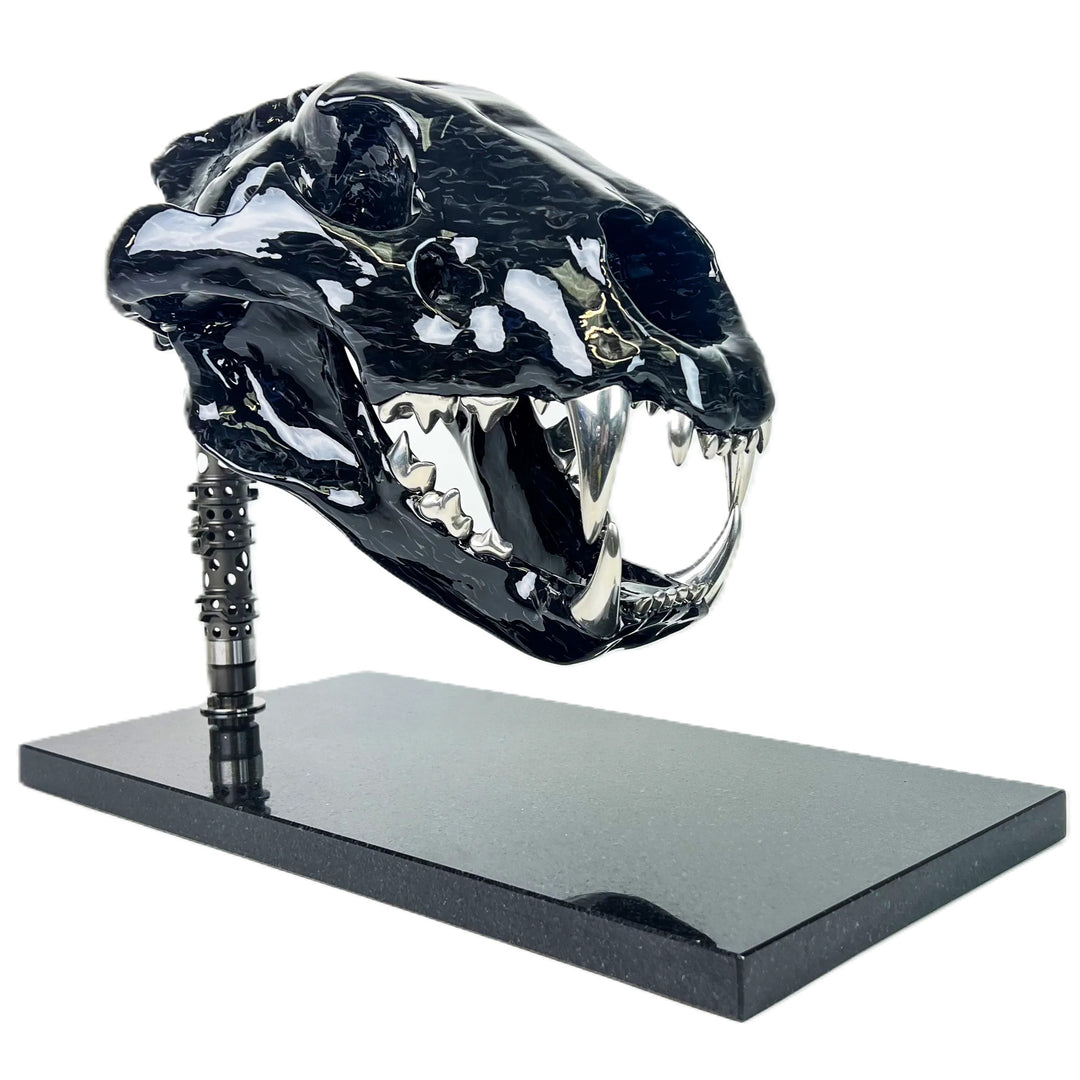 A striking, polished carbon fibre lion skull sculpture by Alastair Gibson, crafted with Mokume-style carbon layup for a swirling, chaotic texture. The jaw is fitted with gleaming bismuth alloy teeth. Mounted on a genuine F1 gearbox selector barrel atop a granite base, the work merges natural form with elite motorsport engineering.