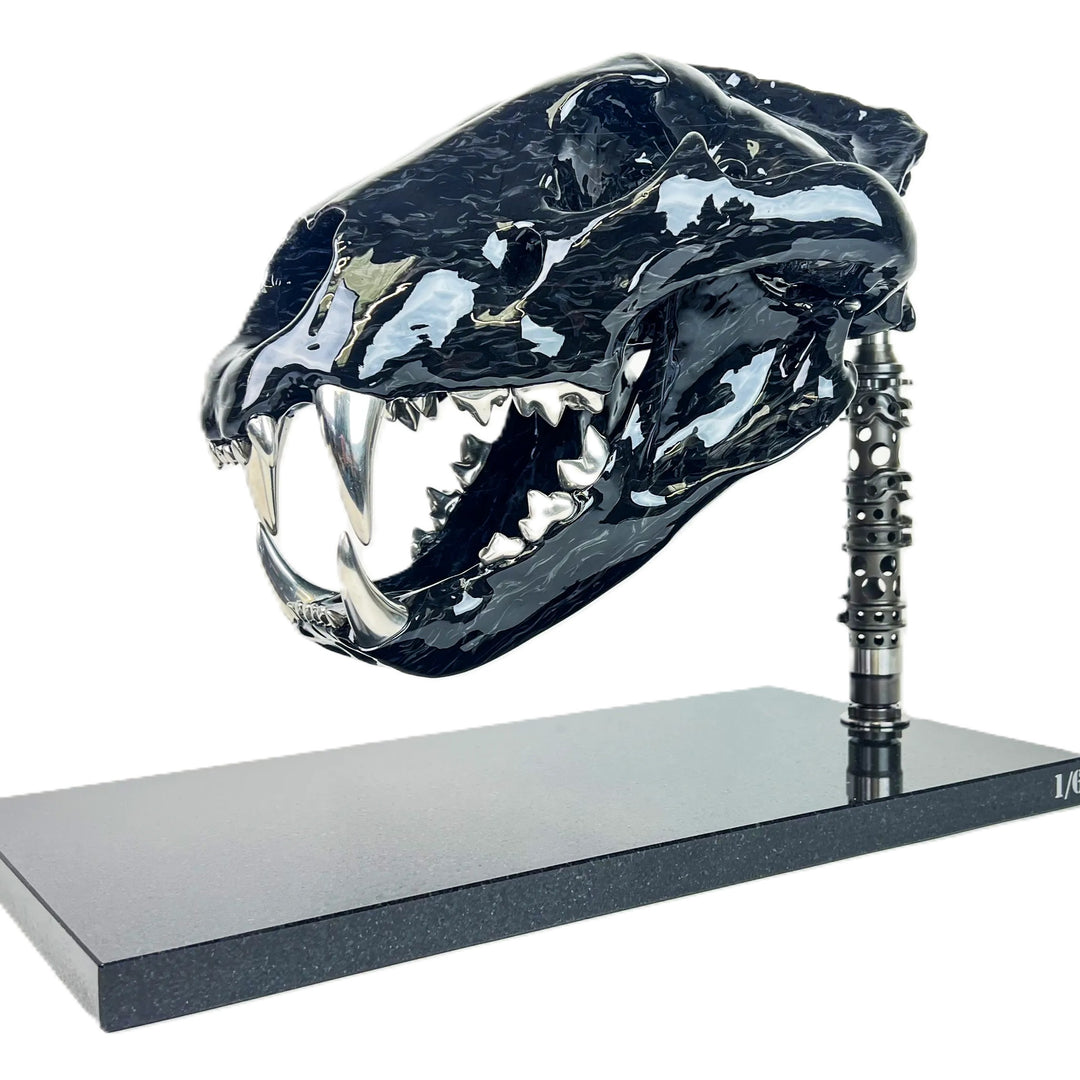 A striking, polished carbon fibre lion skull sculpture by Alastair Gibson, crafted with Mokume-style carbon layup for a swirling, chaotic texture. The jaw is fitted with gleaming bismuth alloy teeth. Mounted on a genuine F1 gearbox selector barrel atop a granite base, the work merges natural form with elite motorsport engineering.