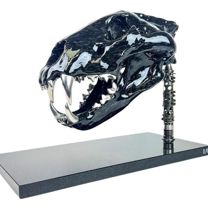 A striking, polished carbon fibre lion skull sculpture by Alastair Gibson, crafted with Mokume-style carbon layup for a swirling, chaotic texture. The jaw is fitted with gleaming bismuth alloy teeth. Mounted on a genuine F1 gearbox selector barrel atop a granite base, the work merges natural form with elite motorsport engineering.