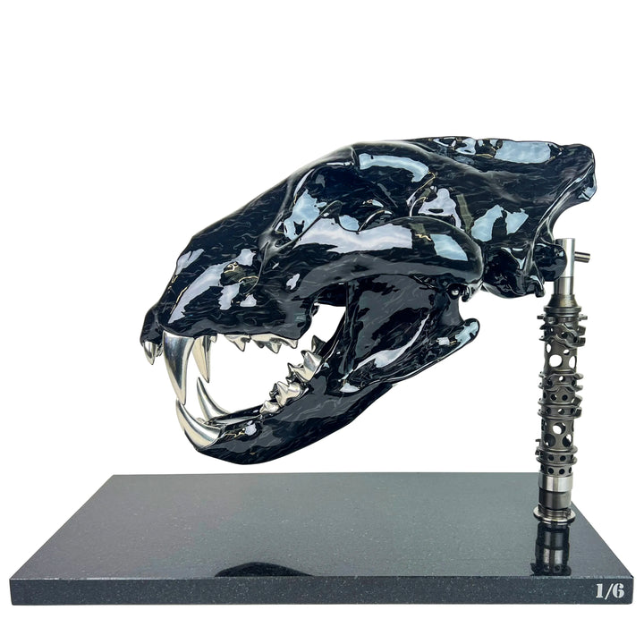 A striking, polished carbon fibre lion skull sculpture by Alastair Gibson, crafted with Mokume-style carbon layup for a swirling, chaotic texture. The jaw is fitted with gleaming bismuth alloy teeth. Mounted on a genuine F1 gearbox selector barrel atop a granite base, the work merges natural form with elite motorsport engineering.