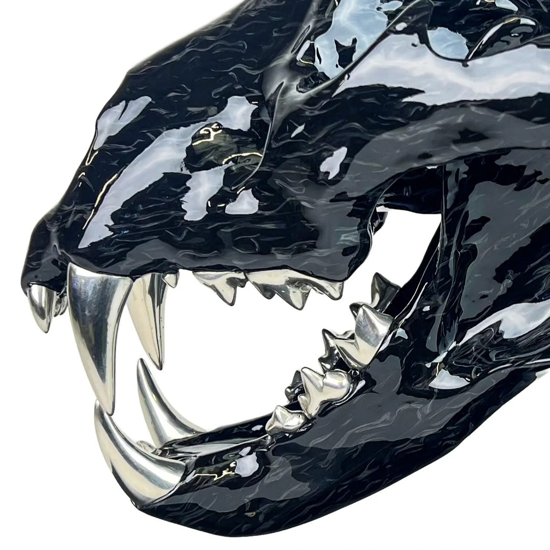 A striking, polished carbon fibre lion skull sculpture by Alastair Gibson, crafted with Mokume-style carbon layup for a swirling, chaotic texture. The jaw is fitted with gleaming bismuth alloy teeth. Mounted on a genuine F1 gearbox selector barrel atop a granite base, the work merges natural form with elite motorsport engineering.