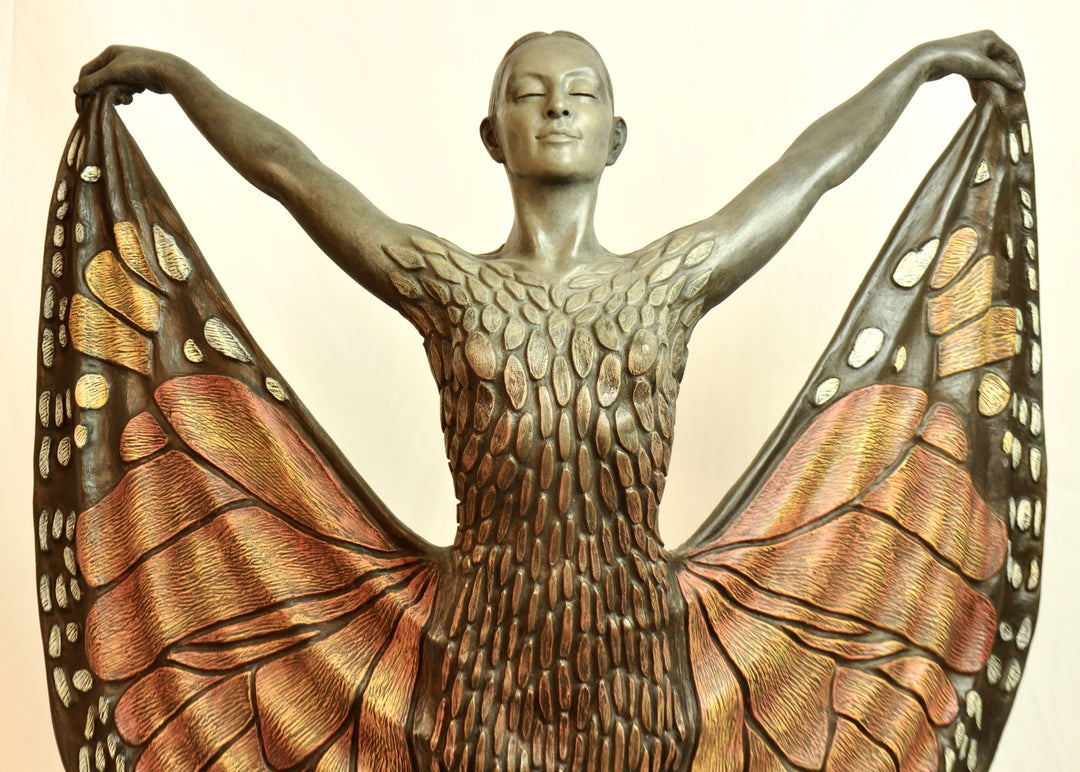 Bronze sculpture of a standing female figure lifting wide, wing-like panels of her dress, revealing intricate monarch butterfly patterns in warm orange, copper, gold and soft cream tones. Her expression is serene and confident, with arms raised and shoulders open as if taking flight. The textured surface and detailed markings mimic butterfly wings while the figure maintains a grounded, elegant posture. The sculpture conveys freedom, transformation and self-expression.