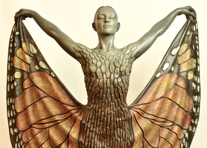 Bronze sculpture of a standing female figure lifting wide, wing-like panels of her dress, revealing intricate monarch butterfly patterns in warm orange, copper, gold and soft cream tones. Her expression is serene and confident, with arms raised and shoulders open as if taking flight. The textured surface and detailed markings mimic butterfly wings while the figure maintains a grounded, elegant posture. The sculpture conveys freedom, transformation and self-expression.