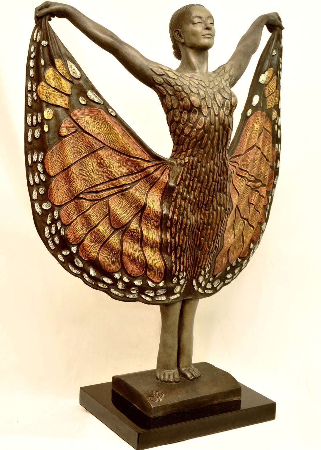 Bronze sculpture of a standing female figure lifting wide, wing-like panels of her dress, revealing intricate monarch butterfly patterns in warm orange, copper, gold and soft cream tones. Her expression is serene and confident, with arms raised and shoulders open as if taking flight. The textured surface and detailed markings mimic butterfly wings while the figure maintains a grounded, elegant posture. The sculpture conveys freedom, transformation and self-expression.