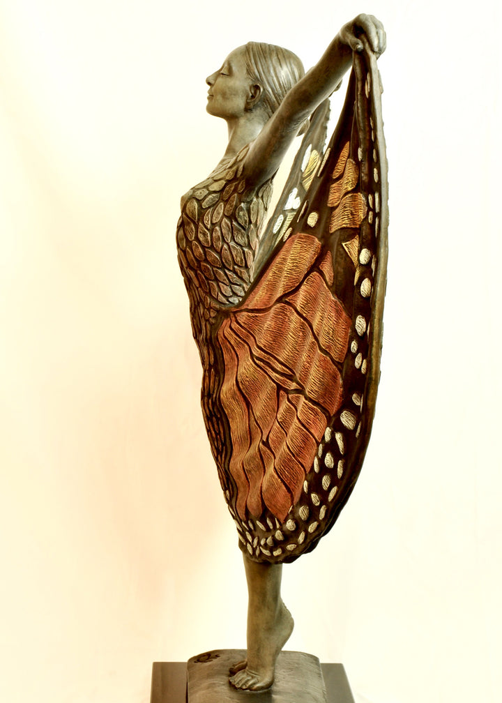 Bronze sculpture of a standing female figure lifting wide, wing-like panels of her dress, revealing intricate monarch butterfly patterns in warm orange, copper, gold and soft cream tones. Her expression is serene and confident, with arms raised and shoulders open as if taking flight. The textured surface and detailed markings mimic butterfly wings while the figure maintains a grounded, elegant posture. The sculpture conveys freedom, transformation and self-expression.