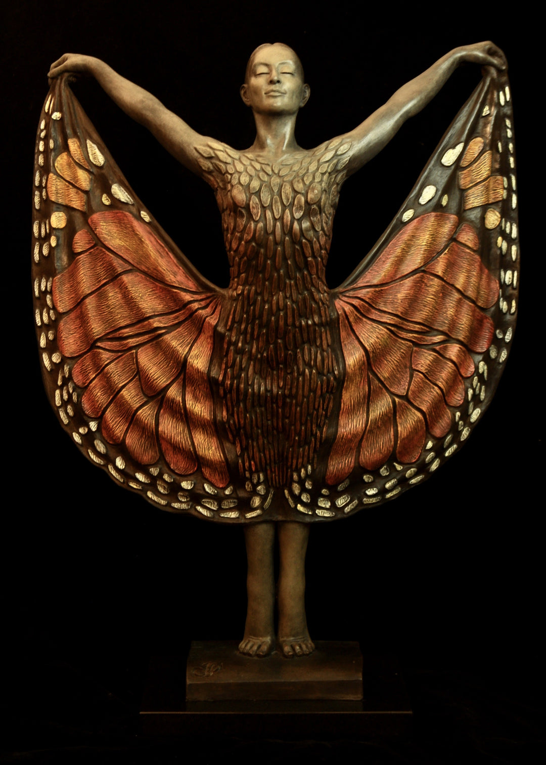 Bronze sculpture of a standing female figure lifting wide, wing-like panels of her dress, revealing intricate monarch butterfly patterns in warm orange, copper, gold and soft cream tones. Her expression is serene and confident, with arms raised and shoulders open as if taking flight. The textured surface and detailed markings mimic butterfly wings while the figure maintains a grounded, elegant posture. The sculpture conveys freedom, transformation and self-expression.