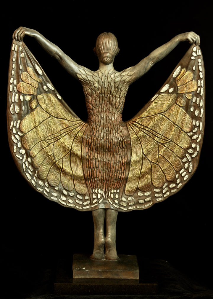 Bronze sculpture of a standing female figure lifting wide, wing-like panels of her dress, revealing intricate monarch butterfly patterns in warm orange, copper, gold and soft cream tones. Her expression is serene and confident, with arms raised and shoulders open as if taking flight. The textured surface and detailed markings mimic butterfly wings while the figure maintains a grounded, elegant posture. The sculpture conveys freedom, transformation and self-expression.