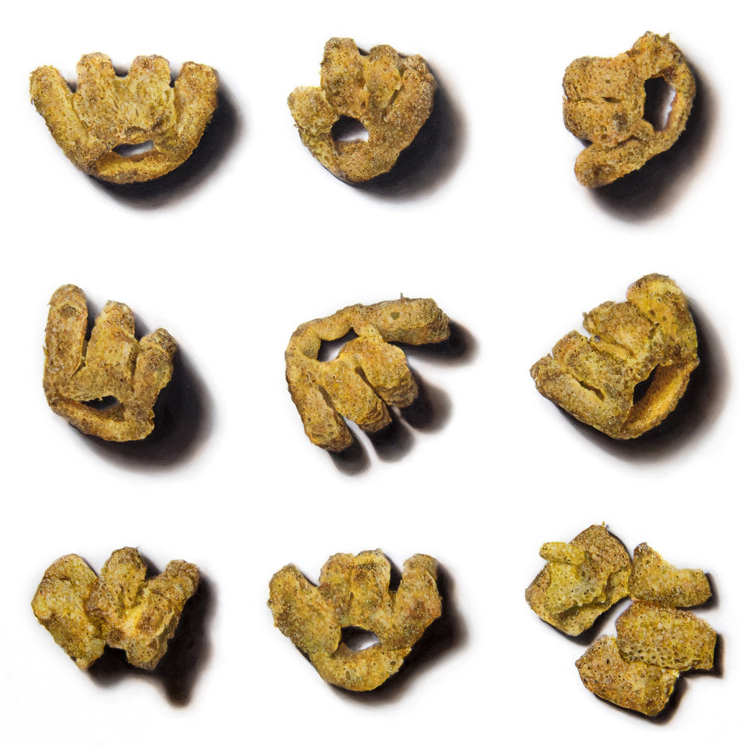 Roast Beef Monster Munch by Simon Taylor – photorealistic oil painting of nine golden, textured crisp snacks arranged in a grid on a white ground, each piece casting soft shadows, celebrating everyday British culture through hyper-detailed wall art.