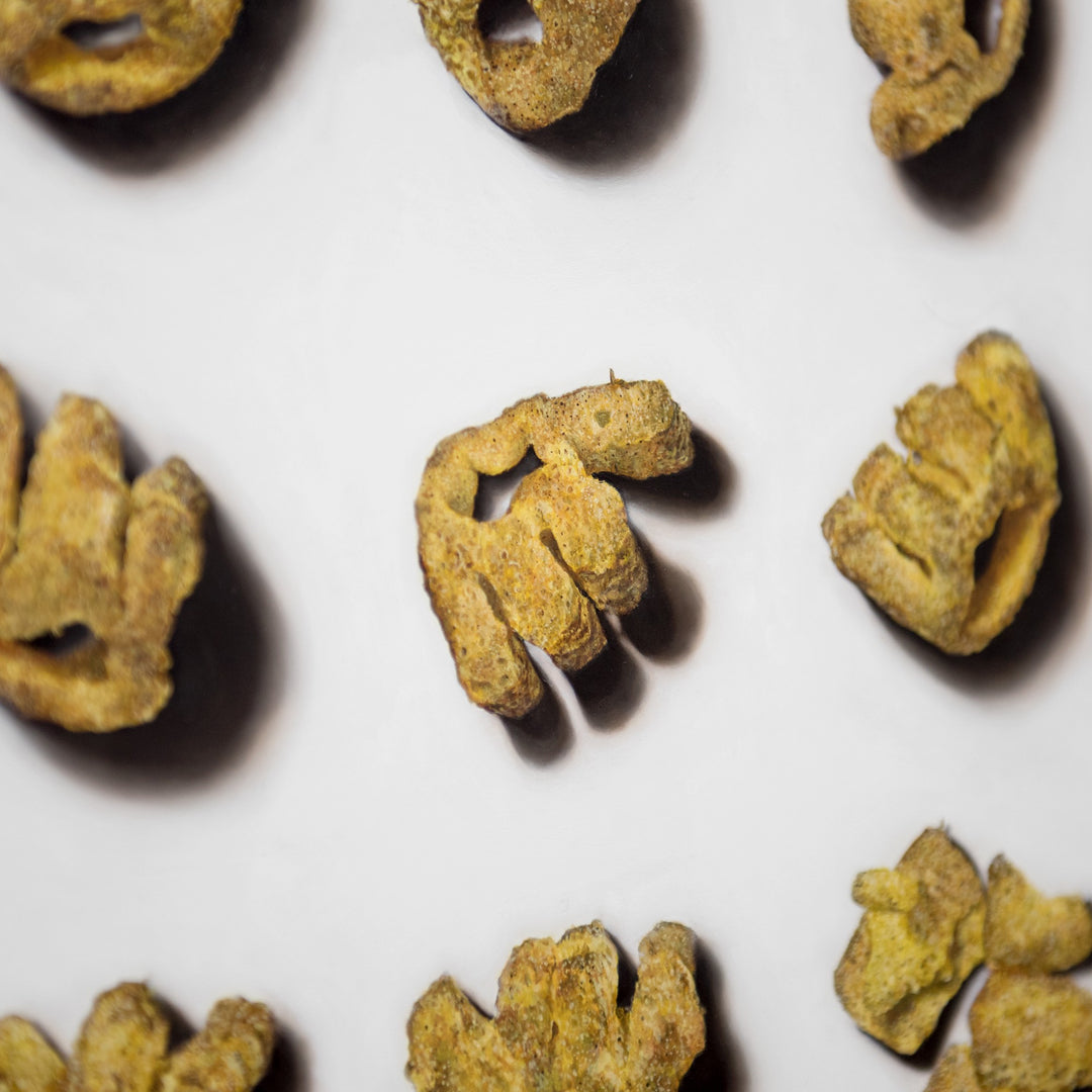 Roast Beef Monster Munch by Simon Taylor – photorealistic oil painting of nine golden, textured crisp snacks arranged in a grid on a white ground, each piece casting soft shadows, celebrating everyday British culture through hyper-detailed wall art.