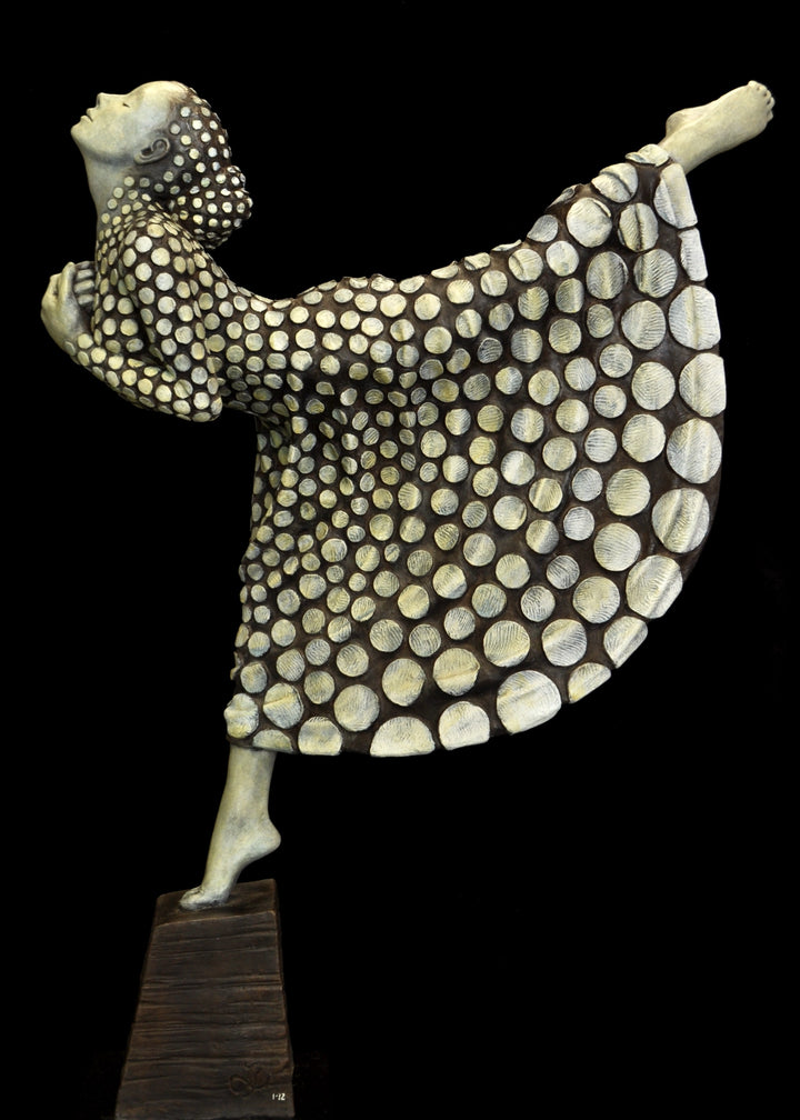 Bronze sculpture of a female figure poised on one leg atop a tiered plinth, her body arched back and arms drawn to her chest as she lifts her face toward the sky. Her dress and skin are patterned with circular, moon-like discs that flow outward into a crescent shape, photographed outdoors in a wide green landscape beneath a cloudy sky.