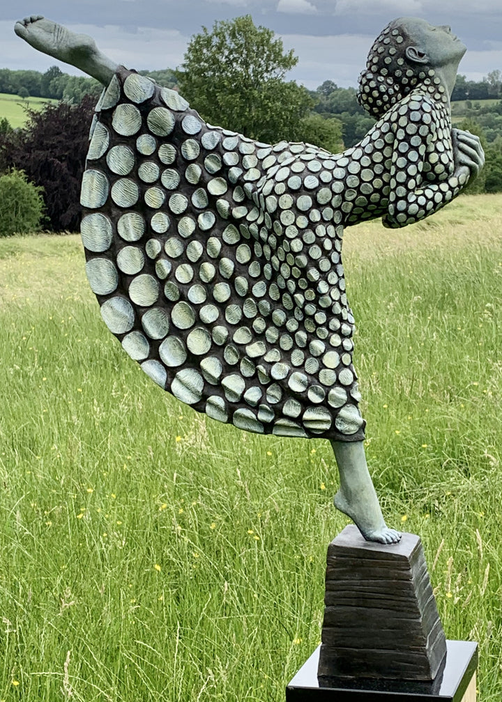 Bronze sculpture of a female figure poised on one leg atop a tiered plinth, her body arched back and arms drawn to her chest as she lifts her face toward the sky. Her dress and skin are patterned with circular, moon-like discs that flow outward into a crescent shape, photographed outdoors in a wide green landscape beneath a cloudy sky.