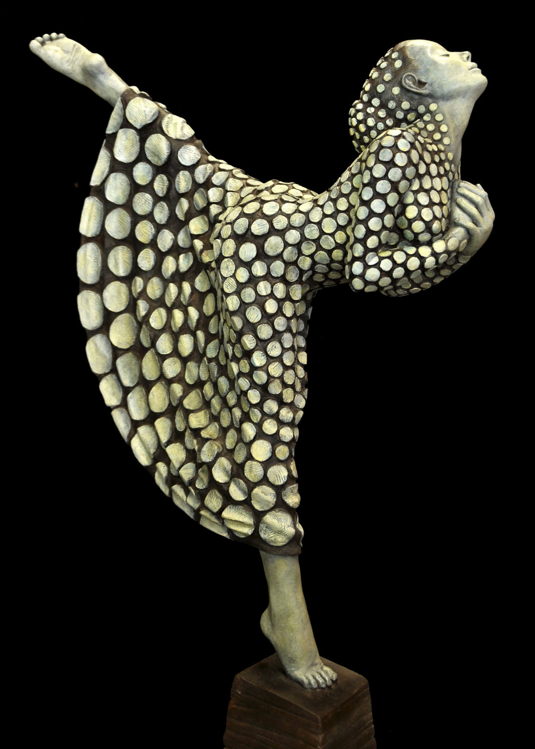 Bronze sculpture of a female figure poised on one leg atop a tiered plinth, her body arched back and arms drawn to her chest as she lifts her face toward the sky. Her dress and skin are patterned with circular, moon-like discs that flow outward into a crescent shape, photographed outdoors in a wide green landscape beneath a cloudy sky.