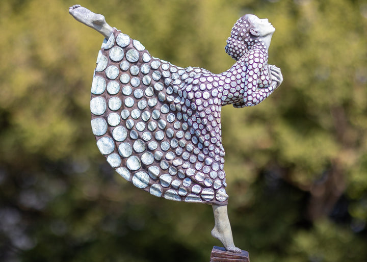 Bronze sculpture of a female figure poised on one leg atop a tiered plinth, her body arched back and arms drawn to her chest as she lifts her face toward the sky. Her dress and skin are patterned with circular, moon-like discs that flow outward into a crescent shape, photographed outdoors in a wide green landscape beneath a cloudy sky.